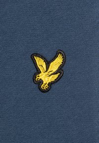 Tissu bleu marine avec un logo d'aigle jaune brodé entouré de noir. La texture est côtelée, ajoutant un détail tactile.