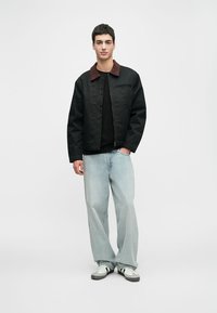 Schwarze Zip-Jacke mit burgunderrotem Kragen, kombiniert mit einem schwarzen T-Shirt und hellblauen weit geschnittenen Jeans. Weiße und schwarze Sneakers.