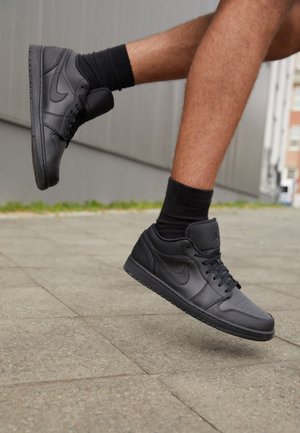 Benen met zwarte leren sneakers en zwarte sokken, één voet iets opgetild boven het trottoir vlak bij de buitenzijde van een gebouw.