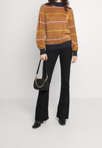 Un pull en bronze et marine à rayures avec une texture brillante, associé à un pantalon évasé noir et un sac à main noir avec un accent en chaîne dorée.
