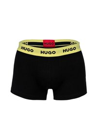 Schwarze Boxershorts mit gelbem Bund, der wiederholt "HUGO"-Logos zeigt, und einem roten Etikett mit dem Schriftzug "HUGO" in der hinteren Mitte.