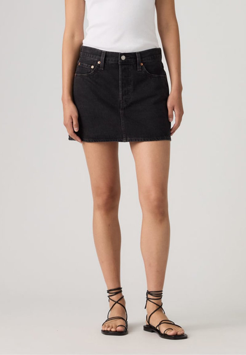 Γυναίκα που φορά μαύρη τζιν μίνι φούστα, λευκό αμάνικο τοπ και μαύρα σανδάλια με λουράκια και επίπεδη σόλα, στέκεται σε απλό ανοιχτόχρωμο φόντο.