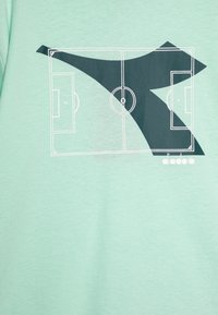 Μίντ πράσινη μπλούζα με λευκό γραφικό ενός γηπέδου ποδοσφαίρου σε σχέδιο, με σκούρες πράσινες αφηρημένες μορφές και το όνομα της μάρκας "diadora" στο κάτω μέρος.