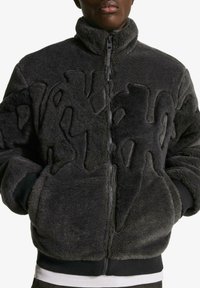 Gray fleece jacket with a high collar, zip closure, and textured abstract design on the front. It features ribbed cuffs and hem.