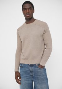 Beżowy sweter z dzianiny z teksturowanymi szwami na ramionach, dopasowany fason i ściągacze przy rękawach, w połączeniu z niebieskimi dżinsami. Gładkie tło.