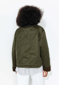 Veste olive avec une texture mate, présentant des poignets marron, une coupe décontractée et un ourlet arrondi, portée sur une chemise rayée.
