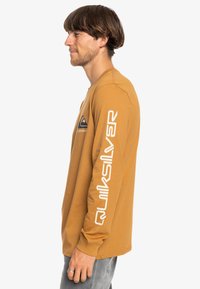 Long-sleeve shirt in mustard color, featuring a blue mountain logo and white text on sleeves. Cotton material, relaxed fit, crew neck design.