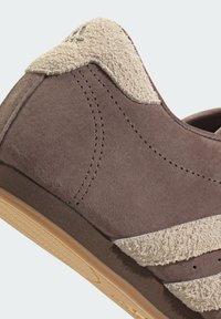 Brown suede sneaker with a textured beige synthetic accent, rounded toe, and a rubber sole featuring a herringbone pattern.