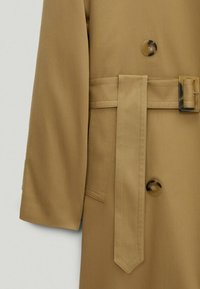Trench-coat beige avec une taille ceintur ée, une fermeture à un seul bouton, des manches longues et une texture de tissu lisse.