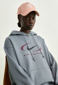 Grauer Hoodie mit einem maronenfarbenen und schwarzen Nike-Logo. Kombiniert mit einer hellrosanen Kappe, die ein weißes Buchstabenlogo zeigt. Glatte Textur.