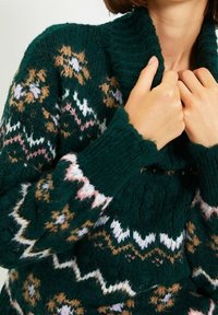 Tummanvihreä neulottu neuletakki, jossa on valkoisia, vaaleanpunaisia ja vaaleanruskeita geometrisiä kuvioita, jota henkilö sovittaa kaulusta lähellä kaulaansa.