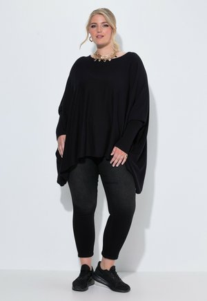 Frau trägt ein lockeres schwarzes Langarmshirt, schwarze Leggings, schwarze Sneaker, eine goldene Halskette und Ohrringe und steht vor einem weißen Hintergrund.