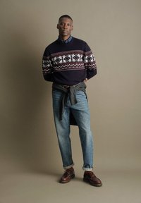 Mann steht vor einfarbigem Hintergrund, trägt gemusterten Pullover, blaue Jeans mit Umschlägen, braune Schuhe und hat eine Jacke um die Taille gebunden.