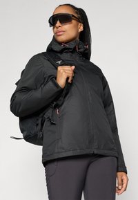 Veste extérieure noire avec un col haut, des poches zippées et des accents rouges. Le modèle porte des lunettes de soleil et porte un sac à dos noir.