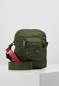 Olīvzaļš neilona plecu soma ar rāvējslēdzēja priekšpuses kabatu, regulējamu auduma siksnu un sarkanu sānos piestiprinātu zīmola birku.