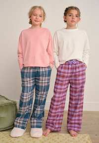 Twee meisjes staan binnen in lange mouwen tops en geruite pyjamabroeken, de een in pantoffels en de ander blootsvoets.