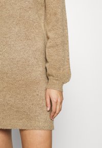 Neulemekko vaaleassa beessissä, jossa on pitkät hihat, ribbatut hihansuut ja pehmeä tekstuuri rentona mallina polvien yläpuolelle.