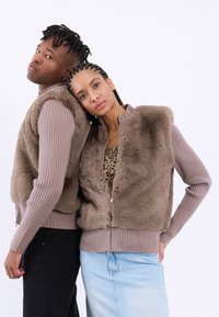 Veste crem din blană artificială cu fermoar, cu mâneci din tricot cu model ridge, având o textură netedă și moale. Un model poartă un top cu imprimeu leopard și o fustă din denim deschis.