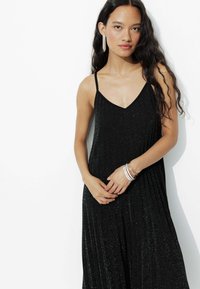 Robe maxi noire sans manches avec un décolleté en V, fabriquée à partir d'un tissu texturé et brillant ; présente un design plissé et des détails superposés.