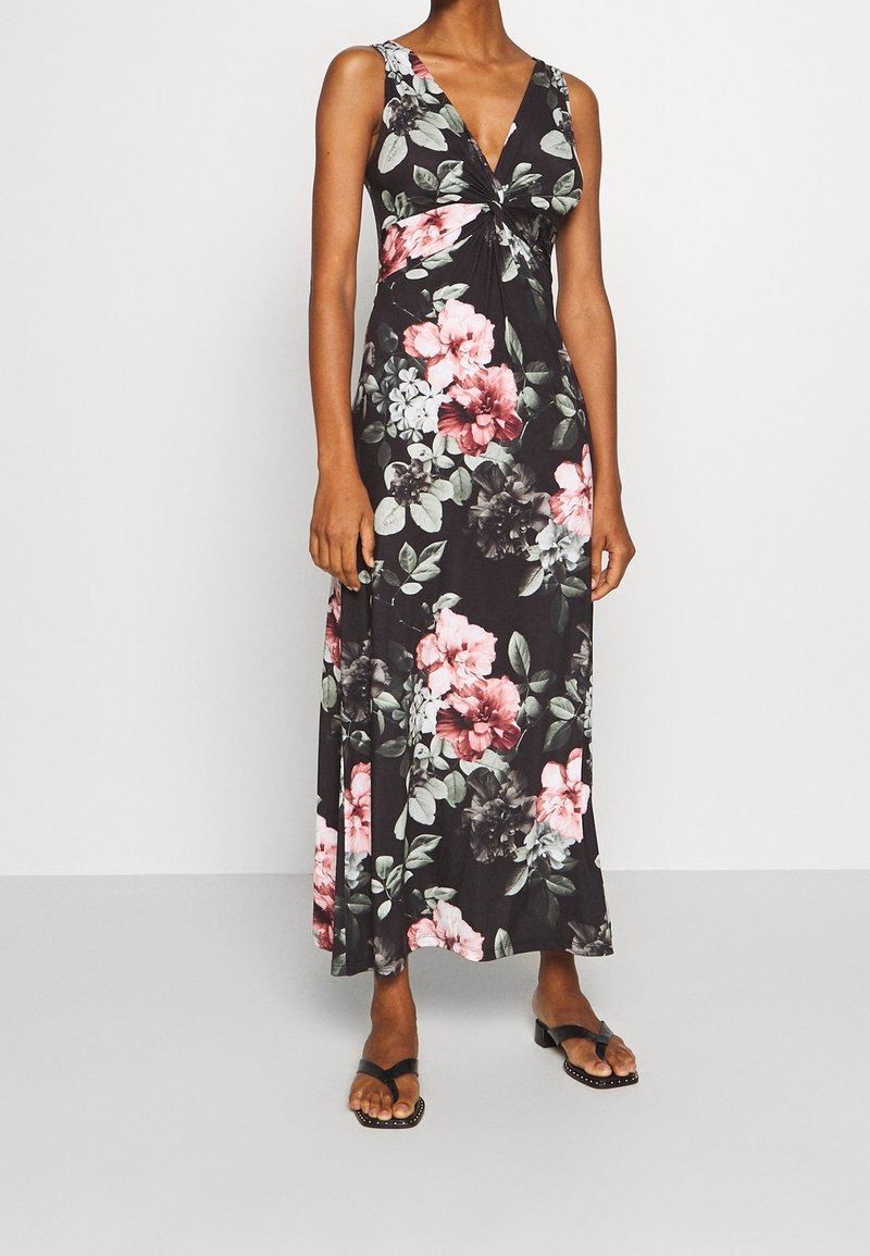 Femme portant une robe longue noire sans manches avec un imprimé floral rose et gris, associée à des sandales noires à bout ouvert à talons bas.