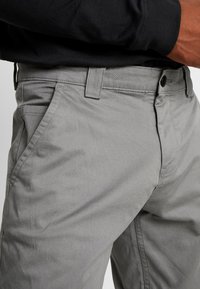 Světle šedé bavlněné kalhoty mají slim fit střih s jemnou texturou, postranními kapsami a pasem s knoflíkovým zapínáním a poutky na opasek.
