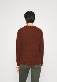 Pull marron texturé avec un col rond et des manches longues, présentant un ourlet arrière légèrement plus long. Porté avec un pantalon vert.