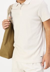 Polo beige clair avec un motif texturé, manches courtes et col. Le modèle tient un sac tote en tissu marron. Design minimaliste.