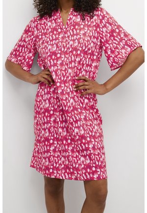 Kobieta ubrana w sukienkę z krótkim rękawem do kolan z abstrakcyjnym, czerwonym, różowym i białym wzorem w plamy, ręce spoczywające na biodrach.