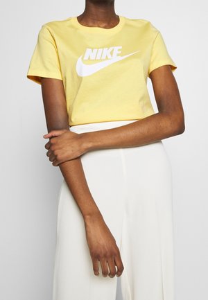 Personne portant un t-shirt Nike jaune avec logo blanc et un pantalon blanc taille haute, bras croisés devant un fond uni.