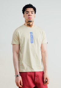 Beige puuvillane T-särk, mille keskel on sinine vertikaalne "ALPHA" graafika. Mudel kannab punaseid pükse ja hõbedast käevõru.