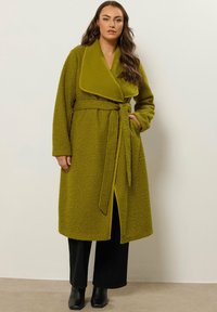 Abrigo de color verde oliva, texturizado, con un amplio cuello, cintura ceñida y bolsillos laterales. Las mangas largas y un dobladillo con abertura completan el diseño.