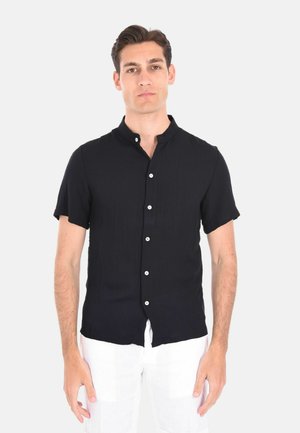 Camicia nera a maniche corte con bottoni in tessuto leggero, colletto band e bottoni bianchi, indossata con pantaloni bianchi.
