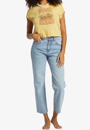 Kobieta w żółtym krótkim T-shircie Billabong i jasnoniebieskich, prostych, jeansach z wysokim stanem, stoi boso z jedną ręką w kieszeni.