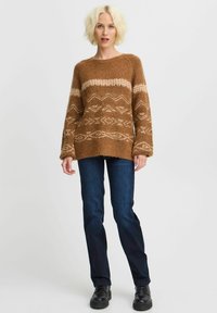 Maglione lavorato a maglia marrone con motivi geometrici color crema, maniche ampie e una vestibilità comoda, abbinato a jeans blu scuro e scarpe nere.