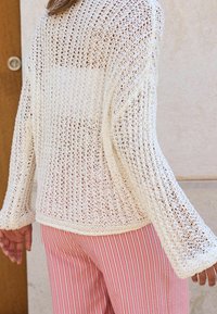 Pull blanc en maille ajourée avec manches cloche, présentant un design ample. Associé à un pantalon rayé rose, sur un fond clair.
