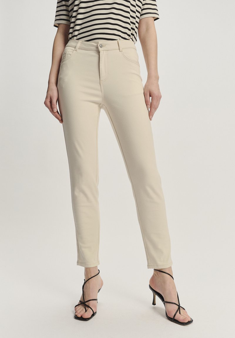 Beżowe dopasowane spodnie z zamkiem błyskawicznym i przednimi kieszeniami. Tkanina mieszanki bawełny z lekkim streczem. Noszone z czarnymi sandałami na wysokim obcasie.