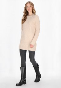 Maglione lavorato a maglia color beige chiaro con scollatura a girocollo e dettagli luccicanti sulle spalle, abbinato a jeans grigi aderenti e stivali neri fino al ginocchio.