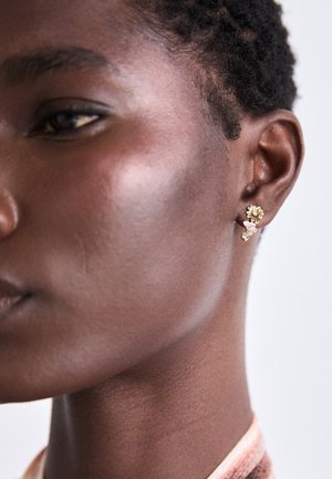 Close-up of dark-skinned person wearing gold flower and pink butterfly drop earrings on the left ear against a plain background.