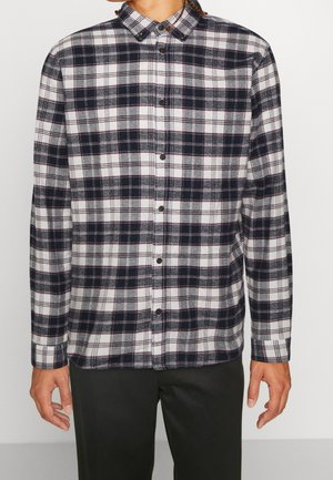 Homme portant une chemise à carreaux noire, blanche et grise à manches longues boutonnée avec un pantalon noir sur un fond uni.