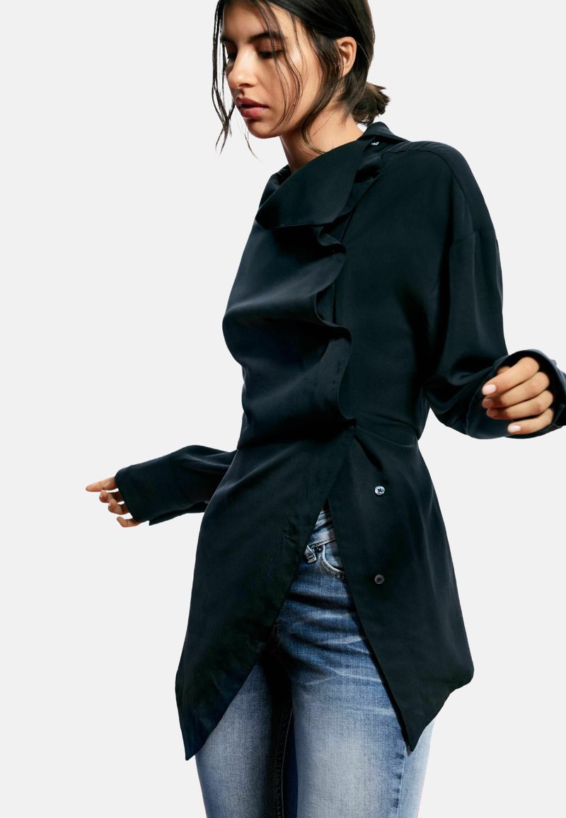 Cămașă neagră cu mânecă lungă și guler înalt, având detalii cu volane și o margine asimetrică. Confecționată dintr-un material neted, cu accente de nasturi.