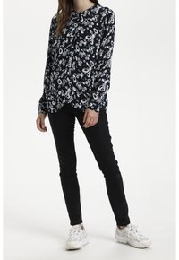 Marinblus med blommönster i vitt och ljusblått, långa ärmar, knäppning framtill, matchad med svarta snäva jeans och vita sneakers.