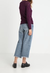 Kobieta w fioletowej bluzce z długim rękawem i koronkowym detalem, w szerokich, ściętych niebieskich dżinsach z postrzępionymi brzegami oraz czarnych butach oksfordkach, stojąca bokiem.