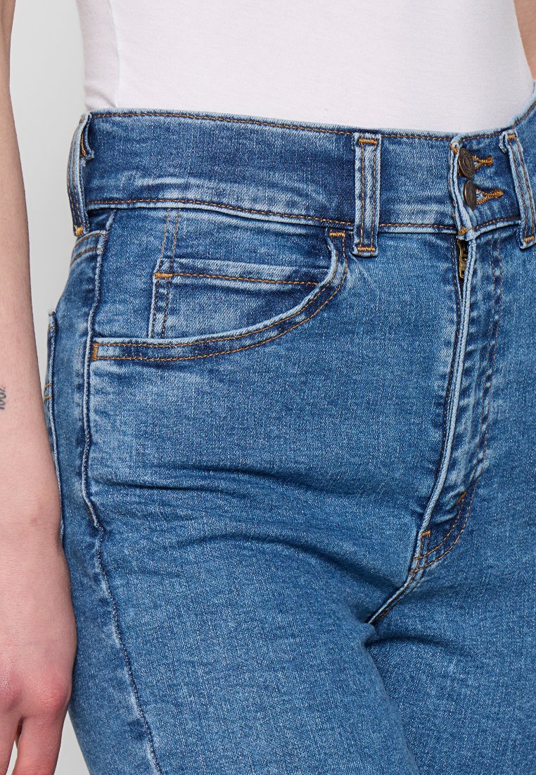 Jeans de denim azul de cintura alta con un ligero efecto de desgaste, costuras naranjas y un diseño clásico de cinco bolsillos. Detalle de cierre con cremallera y botón.