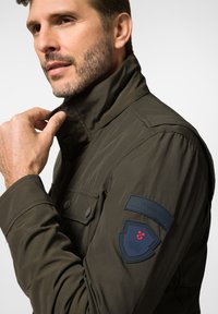 Oliwkowa kurtka z zapięciem pod szyją, o teksturowanej tkaninie i naszywkami na ramionach z logo. Posiada przednie kieszenie i dopasowany krój.