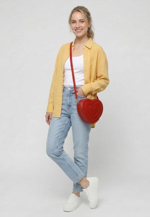 Young woman wearing yellow shirt, white top, light blue jeans, white sneakers, and carrying a red heart-shaped crossbody bag, smiling.