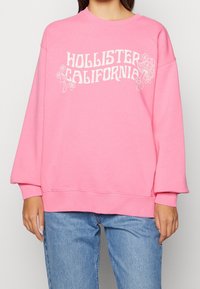 Žena oblečená v růžové mikině s textem "Hollister California" a grafikou hub, v kombinaci s modrými džíny, na jednobarevném pozadí.