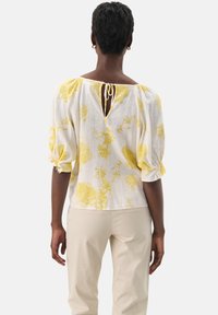 Hvid linnedbluse med gult blomstermønster, puffede ærmer og nøglehulslukning bagpå med snøre. Sæt med beige bukser.