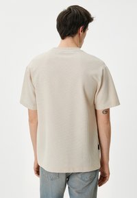 Lyst beige tekstureret T-shirt med korte ærmer, ribbet stof og lige snit. Bagfra ses et lille logomærke forneden.