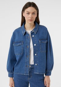 Jeansowa kurtka w średnim odcieniu niebieskiego, luźny krój, z zapięciem na guziki, dwoma kieszeniami na piersi i balonowymi rękawami. Noszona na białej bluzce.