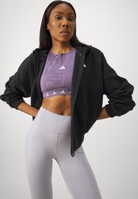 Čierna mikina na zips s šnúrkou, vrstvená nad fialovou športovou podprsenkou s potlačou a svetlosivými legínami s vysokým pásom. Použité textúrované materiály.